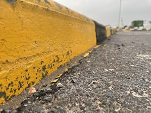 A weathered yellow curb with peeling paint lines the edge of a gritty asphalt road. The ground is speckled with small stones and debris, suggesting a worn, urban environment.