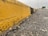 A weathered yellow curb with peeling paint lines the edge of a gritty asphalt road. The ground is speckled with small stones and debris, suggesting a worn, urban environment.