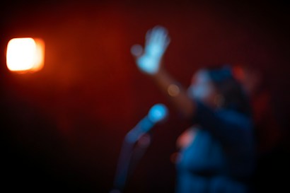 A blurry image depicting a person with an outstretched arm in front of a microphone, set against a dimly lit background with warm orange lighting creating a dramatic effect.