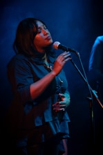A woman is singing passionately into a microphone under soft blue lighting. Her eyes are closed, and she appears deeply immersed in the music. She is wearing a dark blue blouse and has a calm and serene expression.