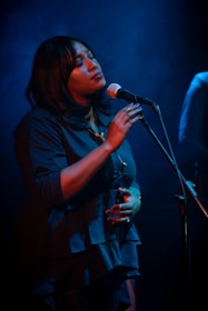 A woman is singing passionately into a microphone under soft blue lighting. Her eyes are closed, and she appears deeply immersed in the music. She is wearing a dark blue blouse and has a calm and serene expression.