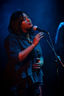 A woman is singing passionately into a microphone under soft blue lighting. Her eyes are closed, and she appears deeply immersed in the music. She is wearing a dark blue blouse and has a calm and serene expression.