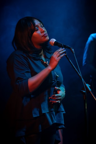 A woman is singing passionately into a microphone under soft blue lighting. Her eyes are closed, and she appears deeply immersed in the music. She is wearing a dark blue blouse and has a calm and serene expression.
