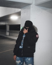 A person wearing a black balaclava and a dark parka jacket with fur trim is standing in a dimly lit parking garage with industrial beams and concrete walls. The person is also wearing ripped blue jeans and a black graphic T-shirt under the jacket. The atmosphere is moody and shadowy, with the background featuring blurry lights and an urban environment.