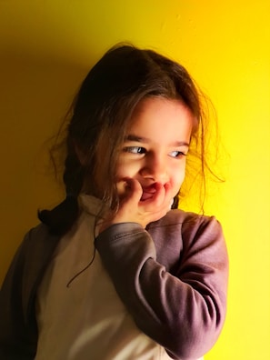 A young child with long, dark hair stands against a vibrant yellow background. The child has a playful expression, with their hand partially covering their mouth, adding a sense of shyness or mischief. The lighting creates soft shadows and highlights, accentuating the warm tones.