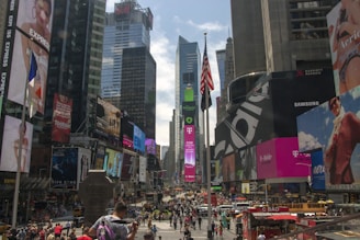 A bustling urban scene with numerous skyscrapers adorned with bright electronic billboards and advertisements, including brands like T-Mobile and Samsung. The American flag is prominently displayed in the foreground, and there is a large crowd of people in the street below, alongside yellow taxis.