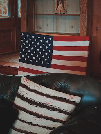 An American flag is draped over a dark green leather couch with a striped pillow. Behind it, there is a wooden cabinet with shelves, and a small figurine or artwork is displayed on one of the shelves. The room has a rustic wooden interior with stained glass on the door.
