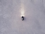 Aerial view of a solitary black car with headlights illuminating the ground, positioned on a vast expanse of textured grayish-white surface.