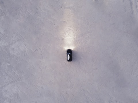 Aerial view of a solitary black car with headlights illuminating the ground, positioned on a vast expanse of textured grayish-white surface.