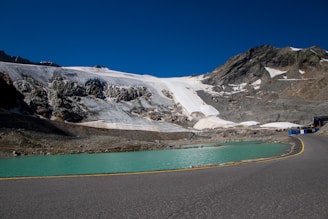 A serene glacier landscape with rugged mountain peaks and a stretch of clear sky above. The foreground features a winding road beside a turquoise glacial lake. Snow and ice patches are visible on the mountainside, indicating chilly conditions.