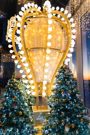 A festive decoration with an oversized, illuminated ornament centerpiece surrounded by Christmas trees adorned with gold and silver baubles and twinkling lights. The scene is set against a backdrop of a window overlooking a sunset.