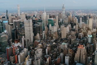 A sprawling city skyline featuring numerous skyscrapers and tall buildings, with New York City as a potential location due to the visible signage. The image captures a mix of modern and classic architecture, densely packed and stretching out toward the horizon under a blue sky.