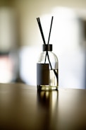 A glass diffuser bottle with a silver top and black reed sticks is placed on a wooden surface. The background is softly blurred, creating a serene atmosphere.