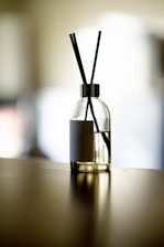 A glass diffuser bottle with a silver top and black reed sticks is placed on a wooden surface. The background is softly blurred, creating a serene atmosphere.
