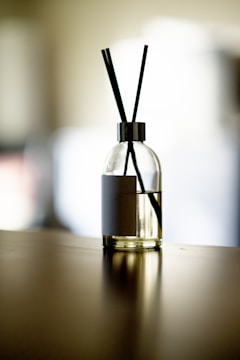 A glass diffuser bottle with a silver top and black reed sticks is placed on a wooden surface. The background is softly blurred, creating a serene atmosphere.