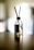 A glass diffuser bottle with a silver top and black reed sticks is placed on a wooden surface. The background is softly blurred, creating a serene atmosphere.