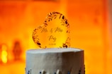 A heart-shaped cake topper made of a transparent material features floral embellishments and the printed text 'Mr. & Mrs. Paz 11/21/20'. It is placed on top of a white frosted cake with blue decorative elements. The background is warmly lit, creating an inviting and celebratory atmosphere.