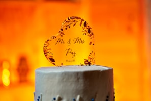 A heart-shaped cake topper made of a transparent material features floral embellishments and the printed text 'Mr. & Mrs. Paz 11/21/20'. It is placed on top of a white frosted cake with blue decorative elements. The background is warmly lit, creating an inviting and celebratory atmosphere.