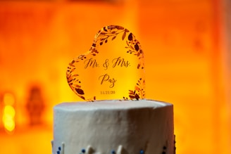 A heart-shaped cake topper made of a transparent material features floral embellishments and the printed text 'Mr. & Mrs. Paz 11/21/20'. It is placed on top of a white frosted cake with blue decorative elements. The background is warmly lit, creating an inviting and celebratory atmosphere.