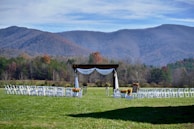 A picturesque outdoor wedding setup is situated in a lush green field. Rows of white chairs are arranged in front of a wooden arbor adorned with white drapery and decorative flowers. Majestic mountains with a hint of fall foliage serve as a breathtaking backdrop under a partly cloudy sky.