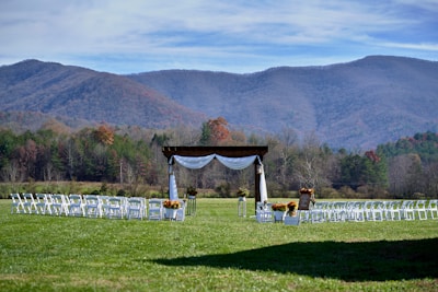 A picturesque outdoor wedding setup is situated in a lush green field. Rows of white chairs are arranged in front of a wooden arbor adorned with white drapery and decorative flowers. Majestic mountains with a hint of fall foliage serve as a breathtaking backdrop under a partly cloudy sky.