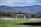 A picturesque outdoor wedding setup is situated in a lush green field. Rows of white chairs are arranged in front of a wooden arbor adorned with white drapery and decorative flowers. Majestic mountains with a hint of fall foliage serve as a breathtaking backdrop under a partly cloudy sky.