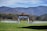 A picturesque outdoor wedding setup is situated in a lush green field. Rows of white chairs are arranged in front of a wooden arbor adorned with white drapery and decorative flowers. Majestic mountains with a hint of fall foliage serve as a breathtaking backdrop under a partly cloudy sky.