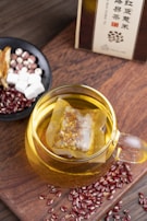A glass cup filled with herbal tea, containing a tea bag immersed in the golden liquid. Surrounding the cup are scattered red beans and a small black bowl containing a mix of ingredients including white cubes, reddish balls, and sliced dried herbs. The setup is placed on a wooden surface, and a box of tea is partially visible in the background.