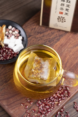 A glass cup filled with herbal tea, containing a tea bag immersed in the golden liquid. Surrounding the cup are scattered red beans and a small black bowl containing a mix of ingredients including white cubes, reddish balls, and sliced dried herbs. The setup is placed on a wooden surface, and a box of tea is partially visible in the background.