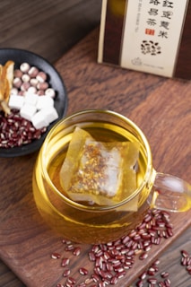A glass cup filled with herbal tea, containing a tea bag immersed in the golden liquid. Surrounding the cup are scattered red beans and a small black bowl containing a mix of ingredients including white cubes, reddish balls, and sliced dried herbs. The setup is placed on a wooden surface, and a box of tea is partially visible in the background.