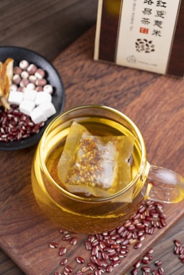 A glass cup filled with herbal tea, containing a tea bag immersed in the golden liquid. Surrounding the cup are scattered red beans and a small black bowl containing a mix of ingredients including white cubes, reddish balls, and sliced dried herbs. The setup is placed on a wooden surface, and a box of tea is partially visible in the background.
