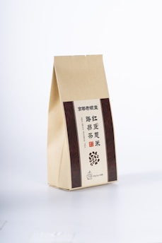 A beige paper package with vertical brown panels on either side, featuring text in Chinese and English. The front includes an illustration of scattered beans and specific details indicating a type of tea.