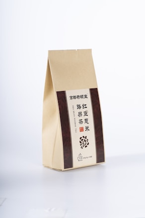 A beige paper package with vertical brown panels on either side, featuring text in Chinese and English. The front includes an illustration of scattered beans and specific details indicating a type of tea.