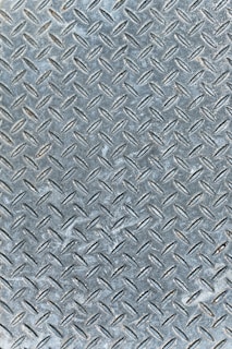 An industrial metal surface with a raised diamond plate pattern, often used for traction and durability. The surface appears weathered with slight discoloration and scuff marks.