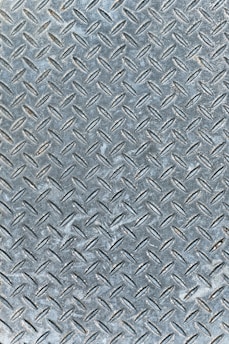 An industrial metal surface with a raised diamond plate pattern, often used for traction and durability. The surface appears weathered with slight discoloration and scuff marks.