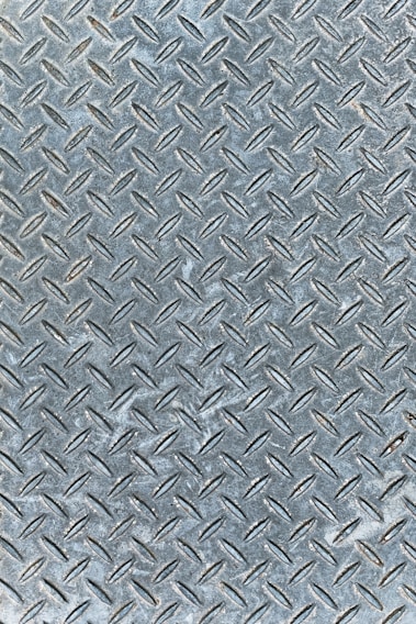 An industrial metal surface with a raised diamond plate pattern, often used for traction and durability. The surface appears weathered with slight discoloration and scuff marks.