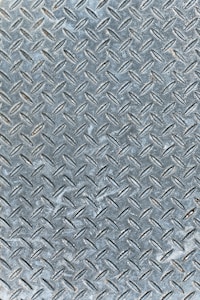 An industrial metal surface with a raised diamond plate pattern, often used for traction and durability. The surface appears weathered with slight discoloration and scuff marks.
