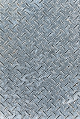 An industrial metal surface with a raised diamond plate pattern, often used for traction and durability. The surface appears weathered with slight discoloration and scuff marks.