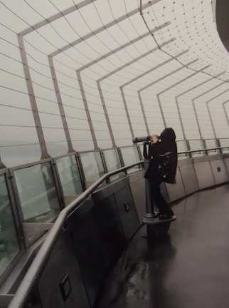 A person in dark clothing is using a telescope on a wet observation deck enclosed by metal railings and mesh. The structure appears to be high up, possibly on a tower, with heavy overcast skies outside.