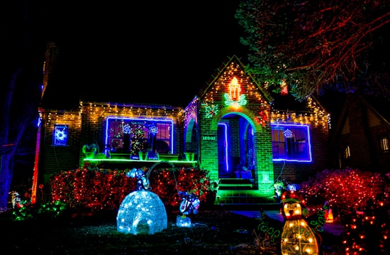 A house is brightly decorated with festive Christmas lights. The exterior features multicolored lights outlining the roof and windows, along with a glowing red wreath on the front door. In the yard, there are illuminated decorations, including a polar bear next to an igloo, small bushes wrapped in red lights, and a light-up snowman.