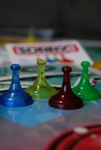 Four translucent game pieces in blue, yellow, red, and green are arranged on a board with a blurred background featuring game cards and other elements.