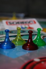 Four translucent game pieces in blue, yellow, red, and green are arranged on a board with a blurred background featuring game cards and other elements.