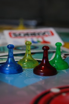 Four translucent game pieces in blue, yellow, red, and green are arranged on a board with a blurred background featuring game cards and other elements.