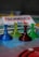 Four translucent game pieces in blue, yellow, red, and green are arranged on a board with a blurred background featuring game cards and other elements.