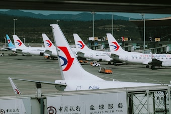Several airplanes are parked at an airport gate, each bearing the China Eastern Airlines logo prominently on the tail. The scene captures a busy airport environment with multiple aircraft, ground service vehicles, and a terminal building in the background. Mountains can be seen in the distance, suggesting an airport located near a hilly or mountainous region.