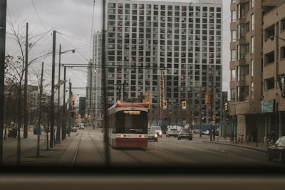 A streetcar is traveling down the center of an urban street lined with high-rise buildings and light traffic. The weather appears overcast, and the streets are relatively calm with few pedestrians visible. Traffic lights hang above the intersection, and the buildings have a modern architectural design.