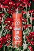 A red candle labeled 'DAME LU' surrounded by vibrant red flowers with green leaves. The design on the candle includes symbols of an eye, flame, and two leaves.
