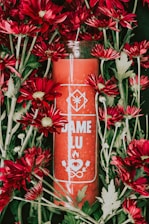A red candle labeled 'DAME LU' surrounded by vibrant red flowers with green leaves. The design on the candle includes symbols of an eye, flame, and two leaves.