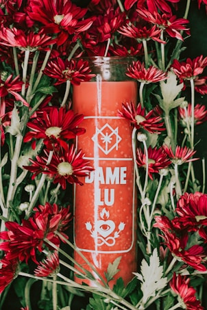A red candle labeled 'DAME LU' surrounded by vibrant red flowers with green leaves. The design on the candle includes symbols of an eye, flame, and two leaves.