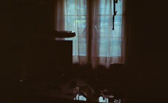 A dimly lit room with sheer curtains covering a window that lets in diffused natural light. The outlines of objects such as a table and a monitor or television are faintly visible. The overall ambiance is dark with a soft, muted glow coming through the window.